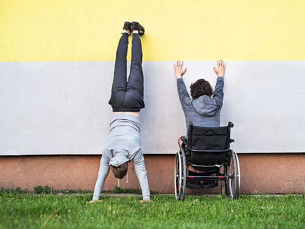 Ein Mensch mit und ein Mensch ohne Behinderung vor einer bunt gestrichenen Hauswand 